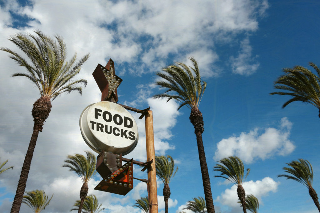 an antique-looking sign surrounded by palm trees that reads "food trucks" with an arrow pointing downward