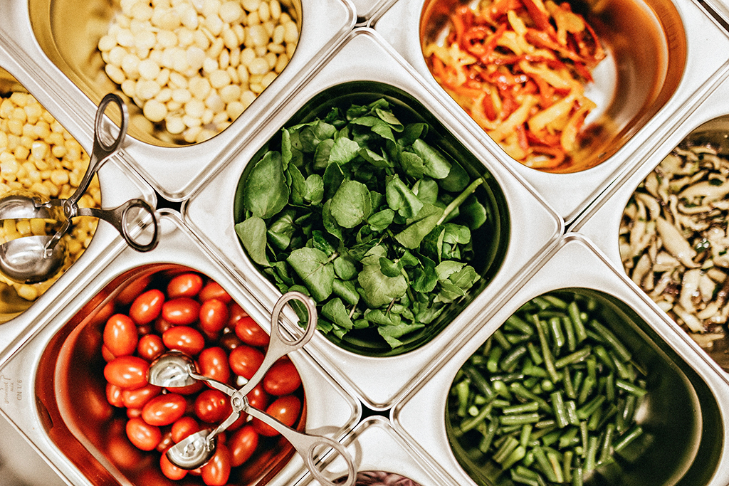 overhead photo of metal sandwich prep food service containers with various vegetables including mushrooms, peppers, tomatoes, corn, green beans and leafy greens