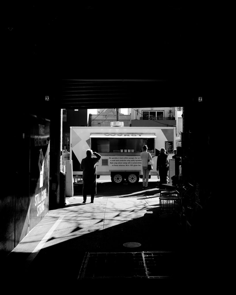 black and white photo looking down a dark alley toward a food truck bathed in light on the street with customers lined up